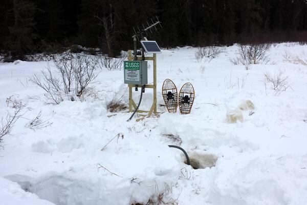 A pair of snowshoes rests in the snow next to a small streamgage mounted on a wooden frame, with antenna and solar panel.