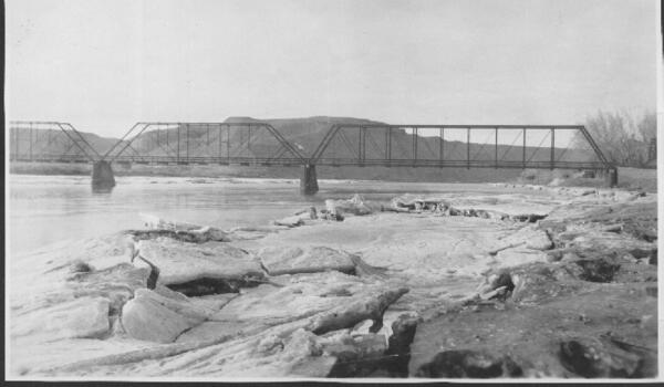 Black and white photo of the old Fort Benton bridge during winter. Ice covers the banks of the river. 