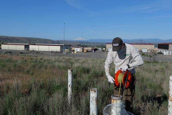 Scientist lowers tape into well in a field with warehouse