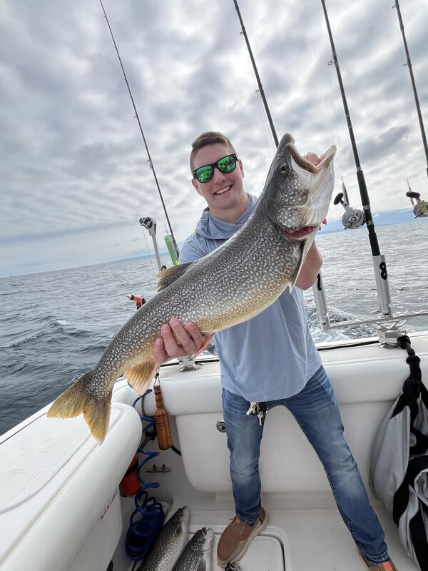 A person on a boat holds up a large fish with speckled coloring and a golden tail. Several fishing rods are positioned upright at the back of the boat, and open water extends into the distance beneath a cloudy sky. 