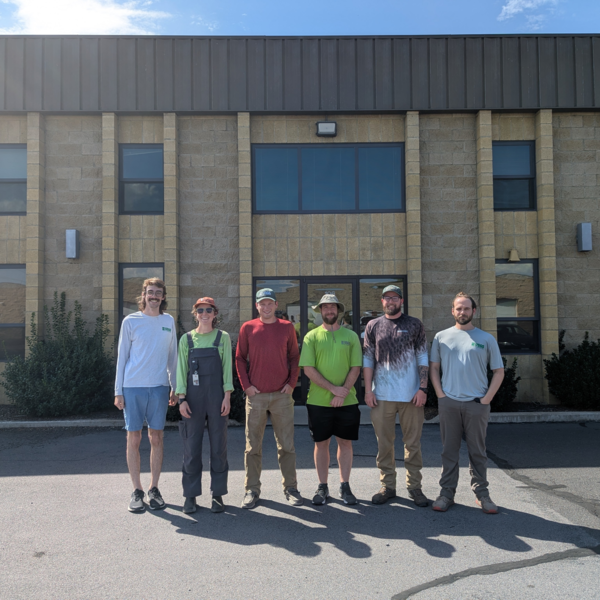 Six people pose in front of a brown brick building on a sunny summer day