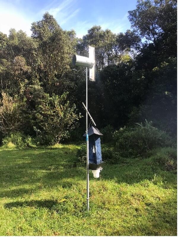 Image shows an aerial spore trap, which is a tall piece of metal with a collector, there are trees and gras in back ground