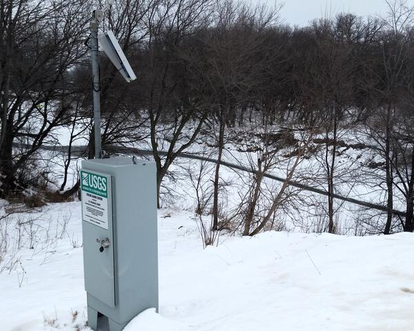 USGS streamgage with solar panel beside a snow-covered riverbank and leafless trees in winter