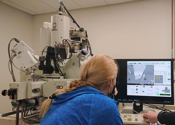 Blonde scientist in a blue hoodie sitting in front of a electron microprobe and computer screen showing a black-and-white image of a crystal