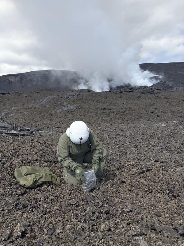 Color photograph of scientist collecting geologic samples near degassing volcanic vent