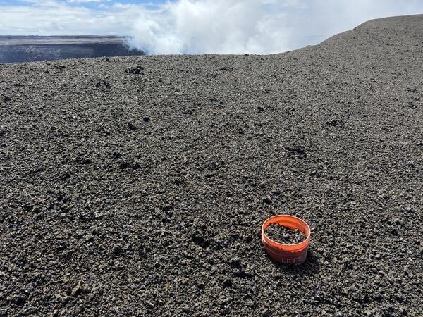 Color photograph of volcanic terrain with bucket in foreground and degassing in background