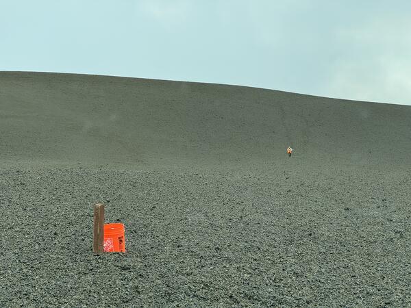 Color photograph of bucket next to mound of volcanic material with person walking in the distance