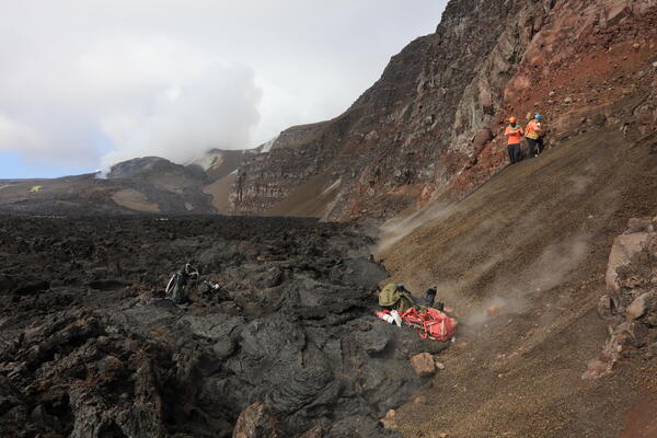 Color photograph of scientists sampling crater wall with volcanic vents degassing in background