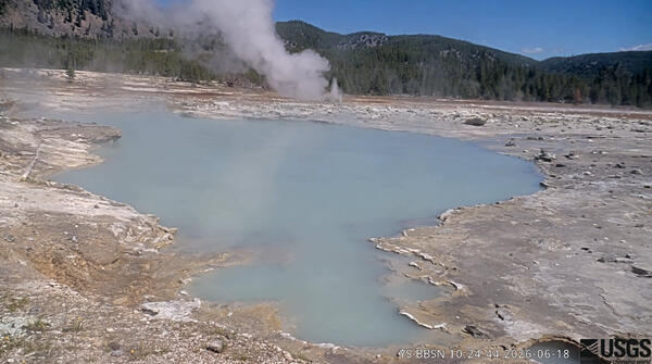 blue pool in a barren area with a treed ridge in the background under blue sky and a small steam vent in the middle ground