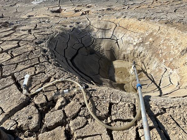 A volcanic gas sampling setup including glass sample bottle connected by tubing to an inverted plastic funnel positioned over a bubbling pool near Salton Buttes, California. A long pole extends from the sampling area, and the surrounding terrain appears dry and fractured.