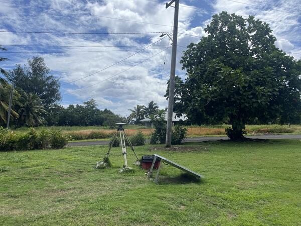 Color photo of a GPS station and solar panel in grass in a tropical setting.