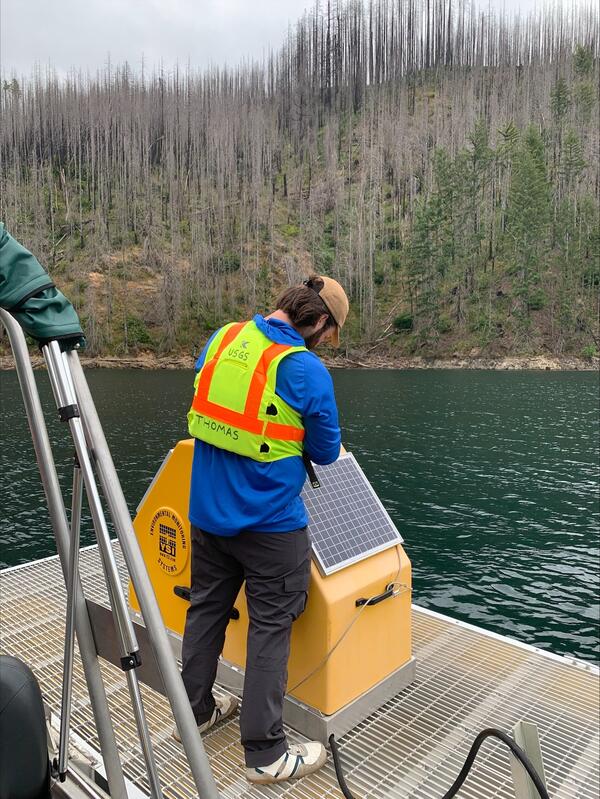 male uses a tool to open gear box on a floating platform in green lake water on a cloudy day. Burned trees in background