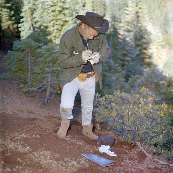 A red-haired man wearing khaki pants, brown boots, a green jacket, and a black cowboy hat stands on a steep, reddish slope surrounded by manzanita bushes and small pine trees. He is taking notes in a small notebook. In the background, a thick pine forest is lit by sunlight.