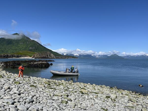 Scientist in orange clothing on rocky shoreline. Skiff in water with male on board with green jacket. Mountains and blue sky.