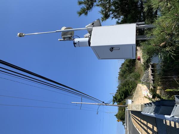 A white square sensor box with solar panel, rain gage, and antenna on the side of a bridge overlooking the water