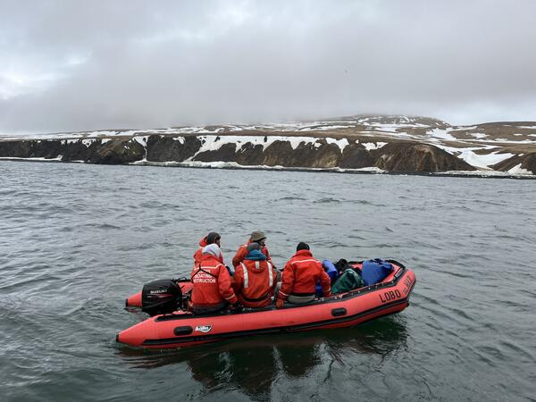 Five scientists wearing orange PFD jackets and hats in skiff with dry bags full of gear. Snow covered cliffs in background.