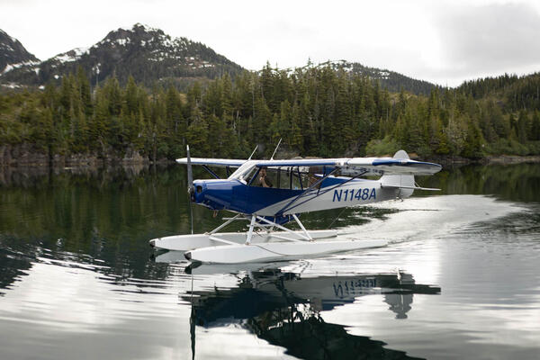 Float plane with pilot giving a thumbs up before takeoff in Prince William Sound. Trees and mountains in background.