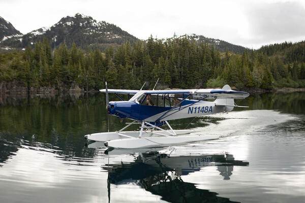 Float plane with pilot giving a thumbs up before takeoff in Prince William Sound. Trees and mountains in background. 