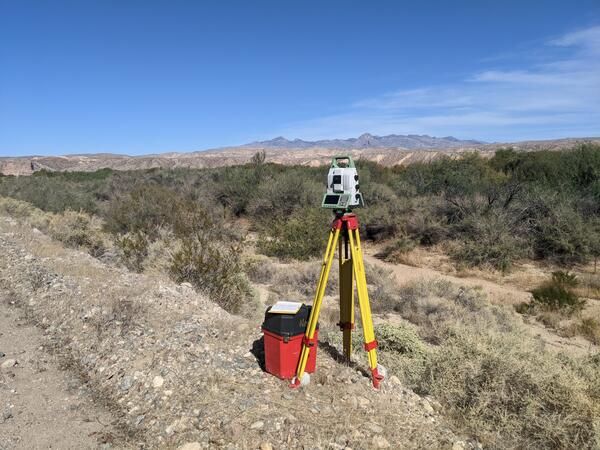 Terrestrial laser scanner on a yellow tripod in a floodplain with brush, distant mountains, and blue sky. 