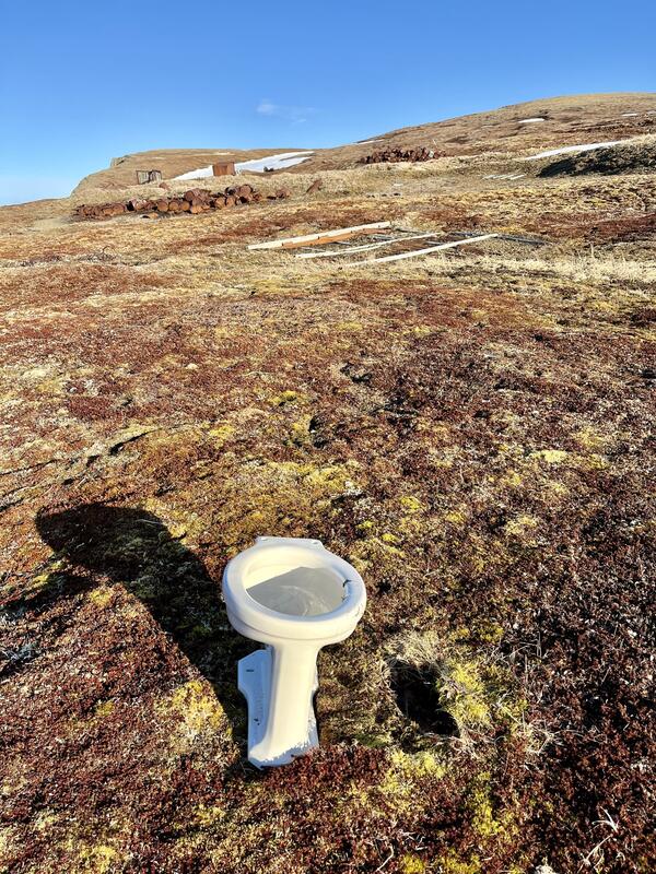 Remains of white toilet on remote island in Alaska. Siting on tundra with artifacts-rusted barrels and wood frames from past.