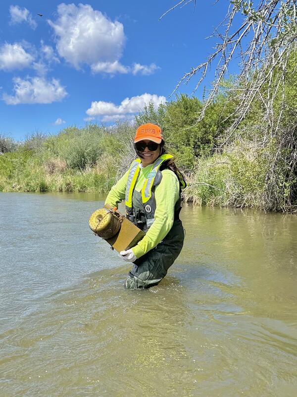 A person wearing waders, PFD, and a USGS hat is standing knee-deep in a river holding a passive pesticides sampler.