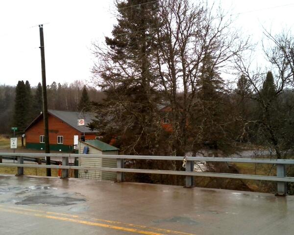 USGS streamgage housing behind bridge guardrails, with a wet road surface on a rural bridge in the foreground.