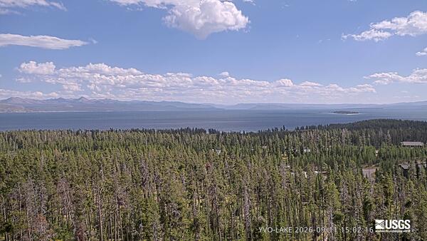 forested area in teh foreground, lake in the middle around, and mountains in the background under partly cloudy sky
