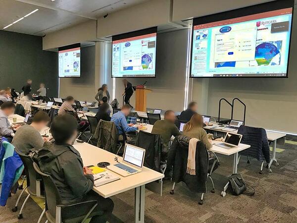 Training room with tables and large screens. Participants sit at the tables.
