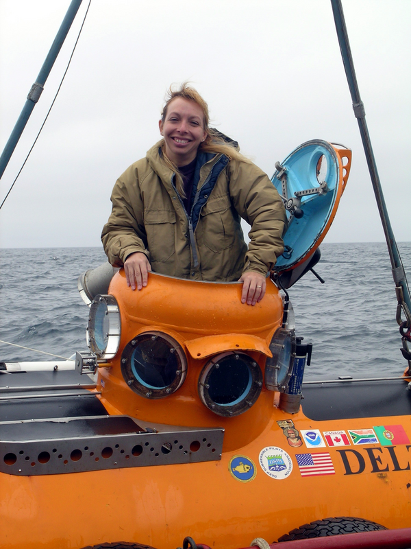 A woman in a brown coat emerges from the hatch of an orange submarine.