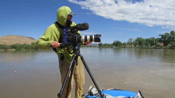 Hooded guy standing on raft with camera on tripod.