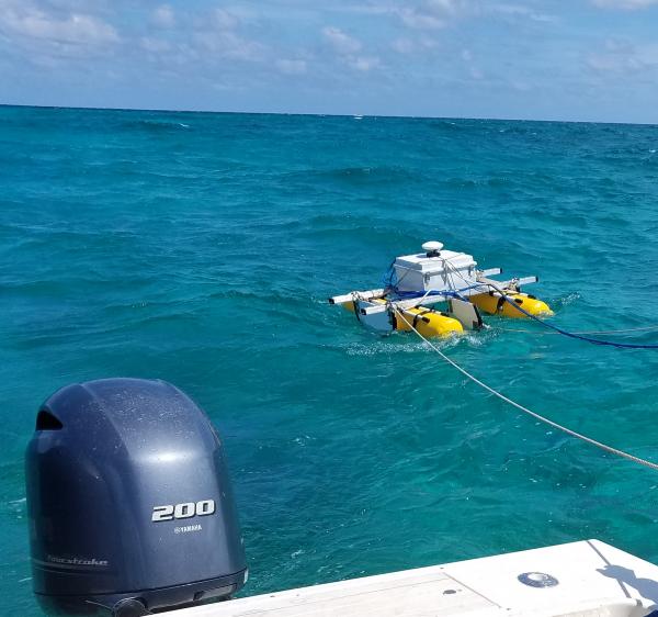 Scientific equipment mounted on two yellow pontoons being towed behind a research vessel over clear blue tropical water