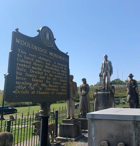 Historical marker installed beside weathered human-figure statues, all enclosed by an iron fence.