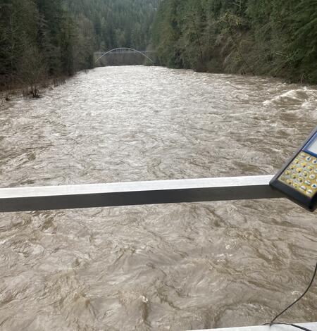 A swollen brown river fills the view. A flow calculator interface is fastened to aluminum rail of cable car.