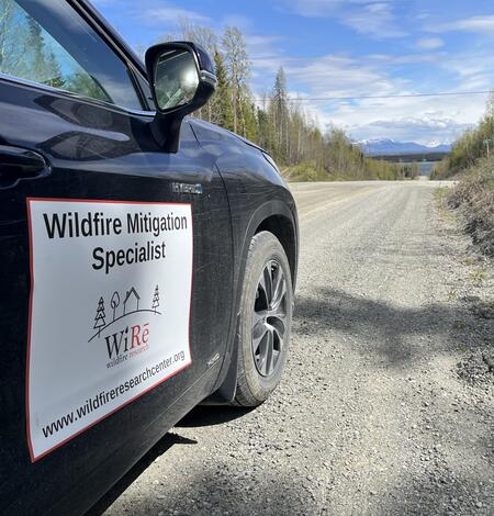 a black car with "wildfire mitigation specialist" on the side drives along a dirt road, through a forest