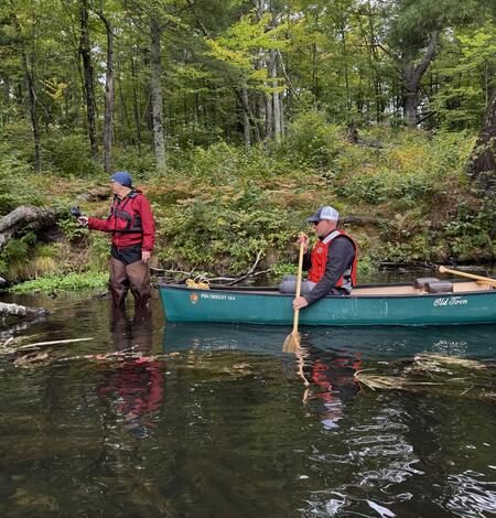 A man in a red vest stands in knee-deep water in a river with a handheld device while another man paddles a canoe.