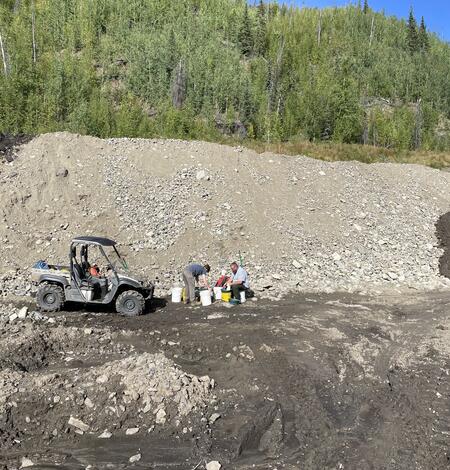 Two scientists next to 4x4 vehicle and buckets. Gravel-rock hill, tire tracks in mud, green trees, blue sky in background.