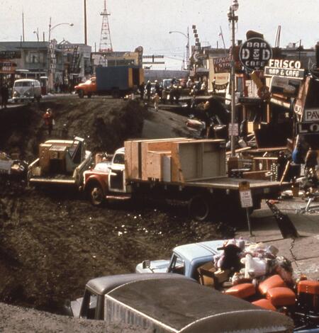 Downtown Anchorage street has sunk now holding several large trucks. Buildings and signs on right side damaged. 