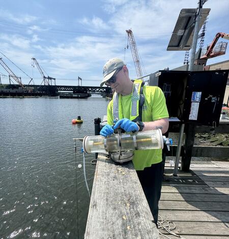 A man in a cap, neon yellow shirt, and blue gloves holds a Van Dorn sampler on a dock with a streamgage and bridge behind.