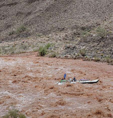 A research boat navigates high rapids in the brown sediment-filled Colorado River during a storm