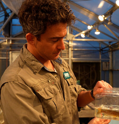 A man with curly hair stands in a laboratory filled with large fish tanks and holds a jar with two small fish swimming