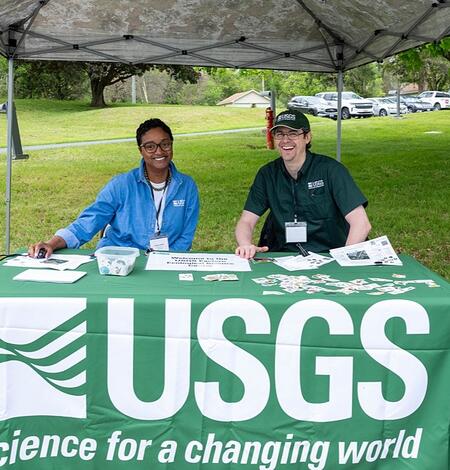 Two USGS employees smile while sitting behind a USGS tablecloth covered table under a pop up tent