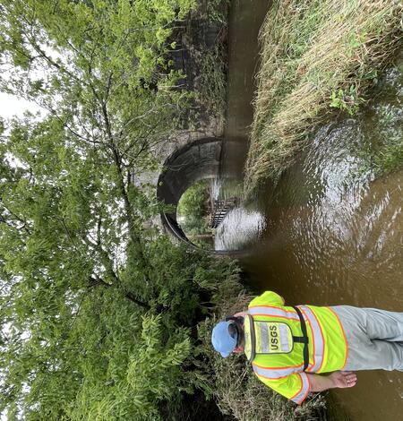 A USGS technician in a bright green safety jacket takes a photo of a flooded river