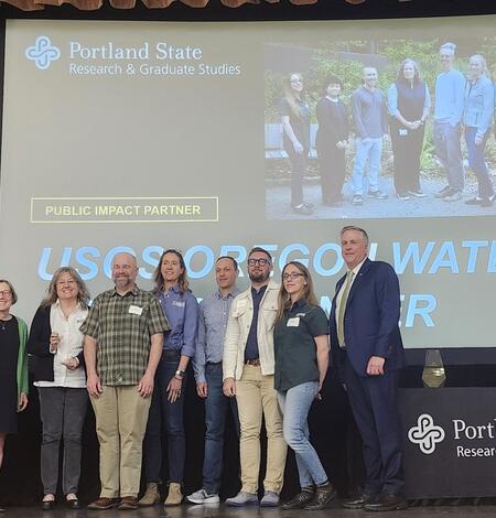 8 people stand on a stage in front of a projection screen with award info on it