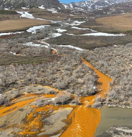 Orange colored water flowing into Kugororuk River, Alaska. Brown ground with vegetation, snow patches with mountains sky.