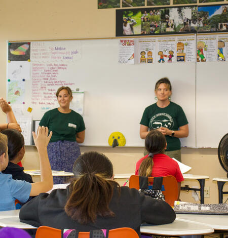 Two people stand at the front of a classroom of children. 