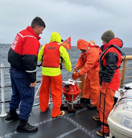 Four people on ship in ocean, huddled around a piece of equipment near railing