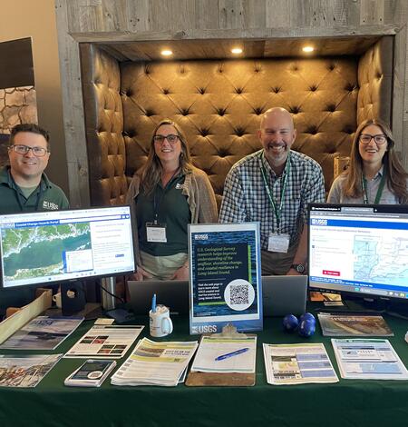 Group of USGS scientists smiling for photo behind booth with handouts and two computer monitors displaying maps
