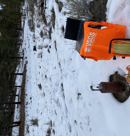 Electronic measuring tape next to a private well cap on a snow dusted landscape with evergreens in the distance
