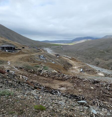 Old buildings, rusted barrel, rocks, and scattered wood from previous buildings in mountain valley. Helicopter near stream.