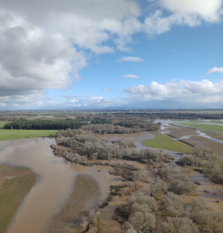 Sediment filled brown meandering river floods into nearby agricultural land. Partly cloudy day.